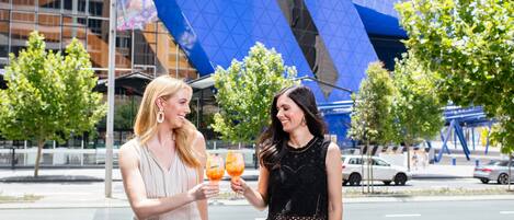 Two women toasting with drinks in front of a modern building with a blue glass facade.