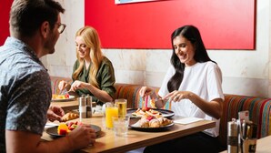A group of people dining at a restaurant with a red wall and a colorful striped bench.