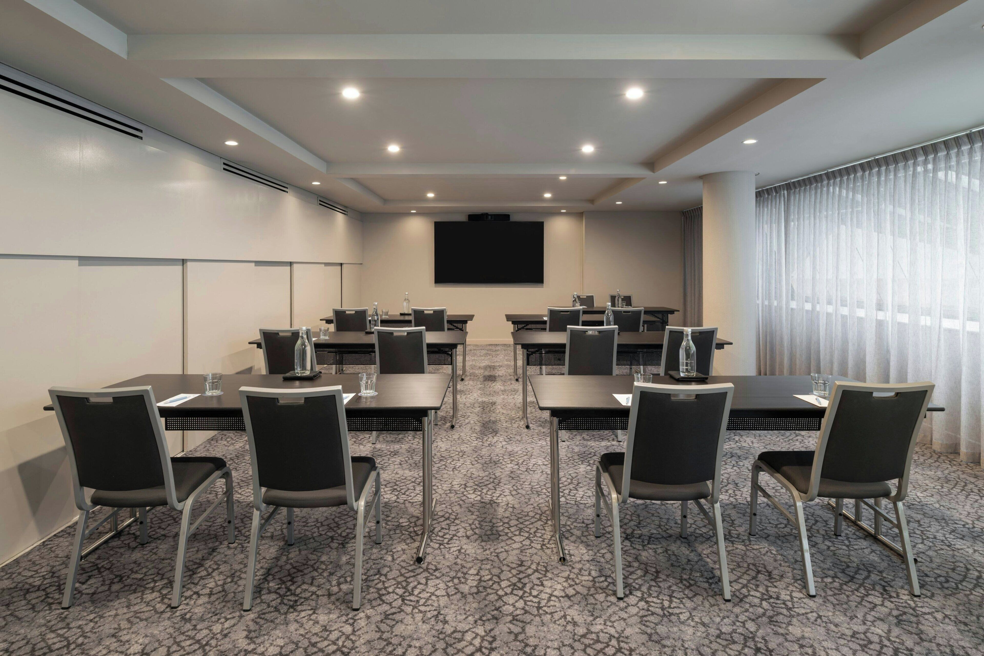 Un salón de conferencias con mesas, sillas, una pantalla de proyector y un amplio ventanal con cortinas.