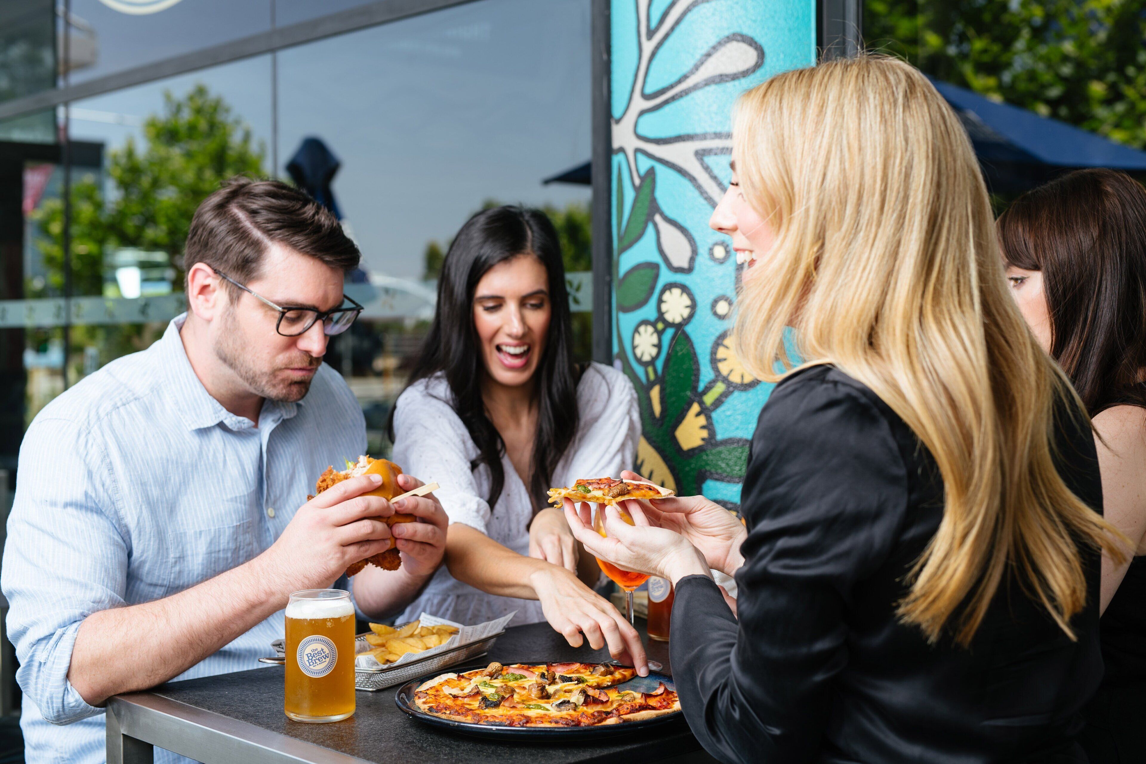 Un groupe de personnes est assis à la terrasse d’un restaurant, où elles dégustent des pizzas et boivent de la bière.