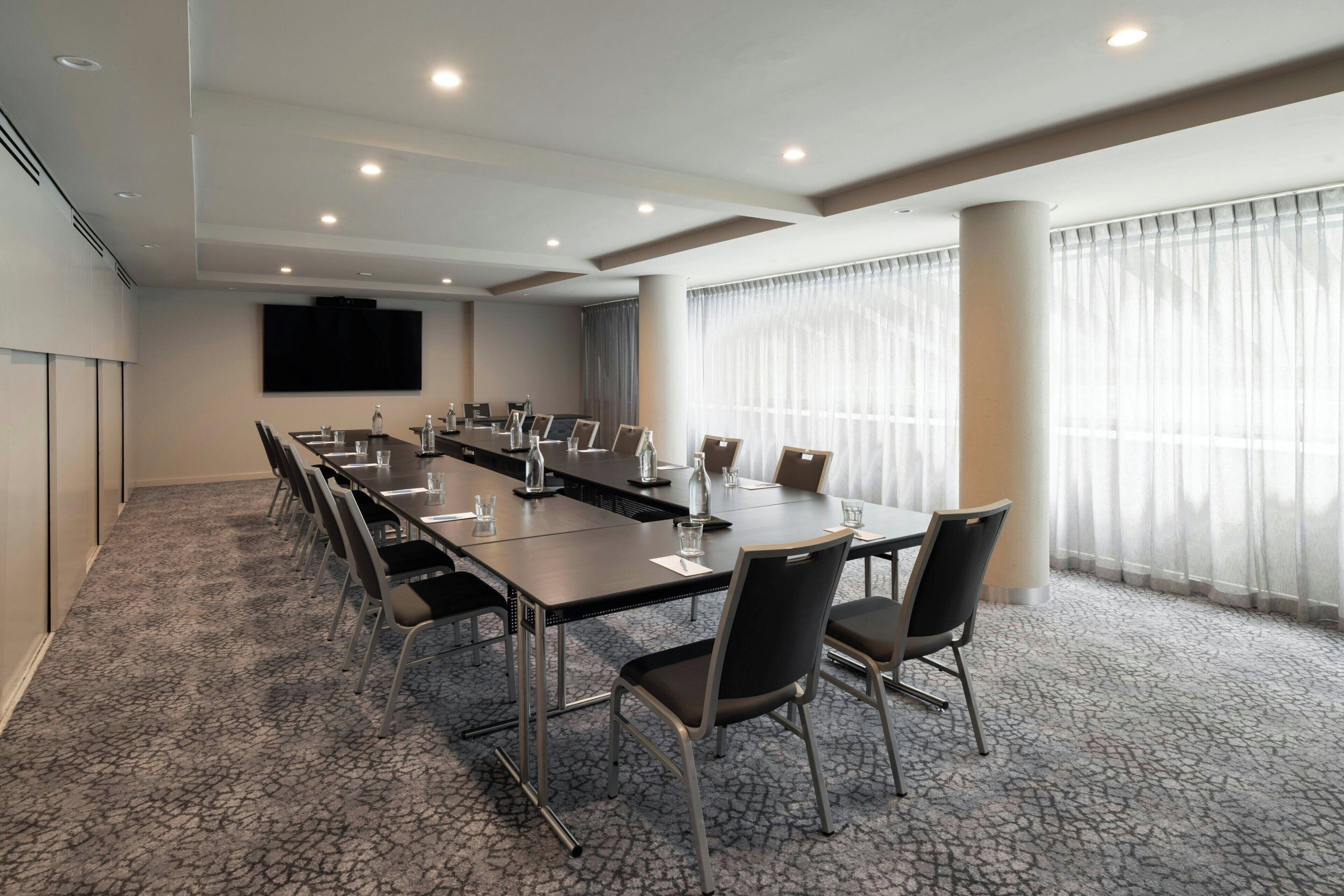 A conference room with a long table, chairs, a screen, and a large window with curtains.
