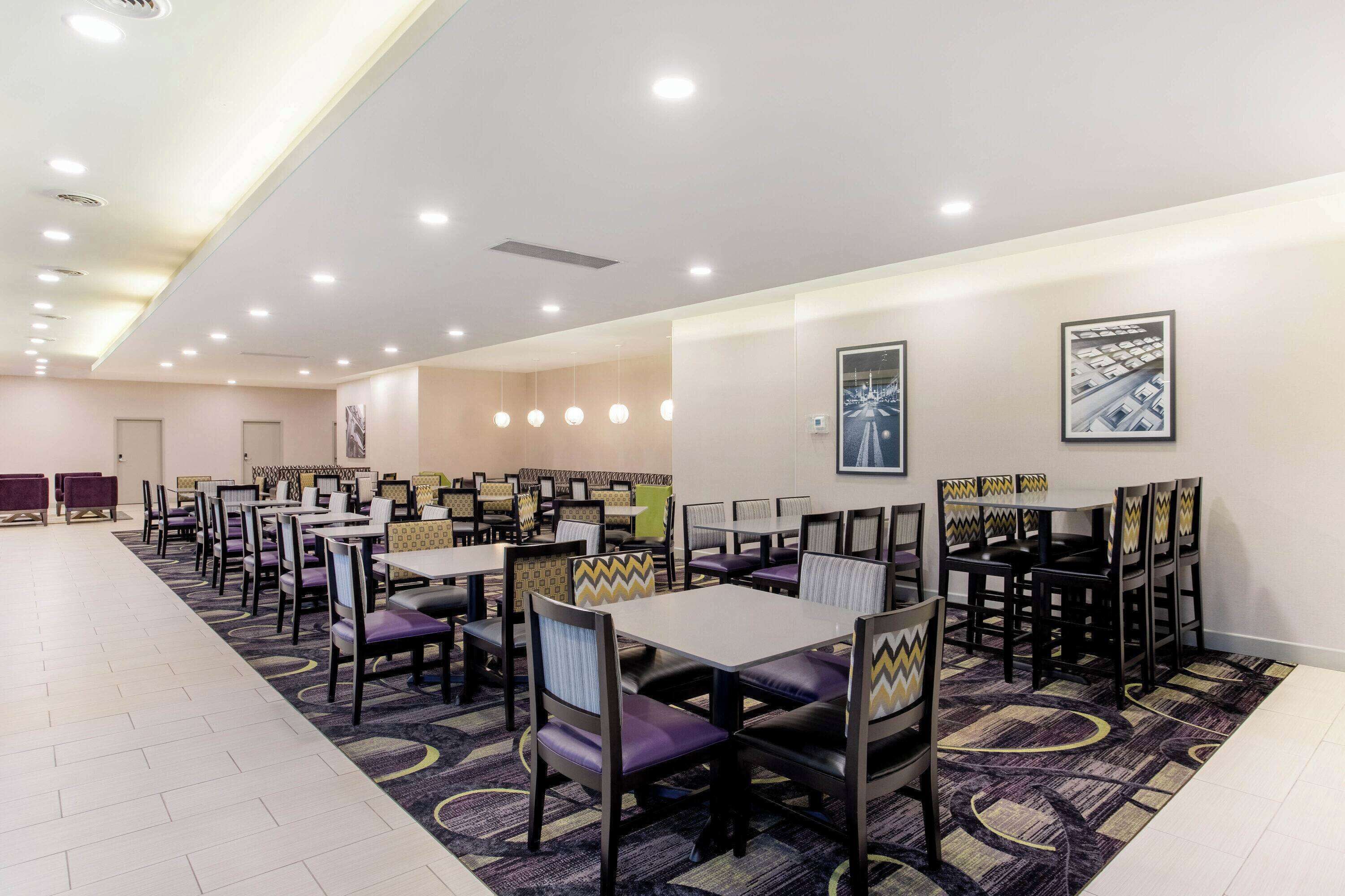 Un comedor de hotel con varias mesas y sillas dispuestas en filas, con luces de techo e imágenes colgadas en las paredes.
