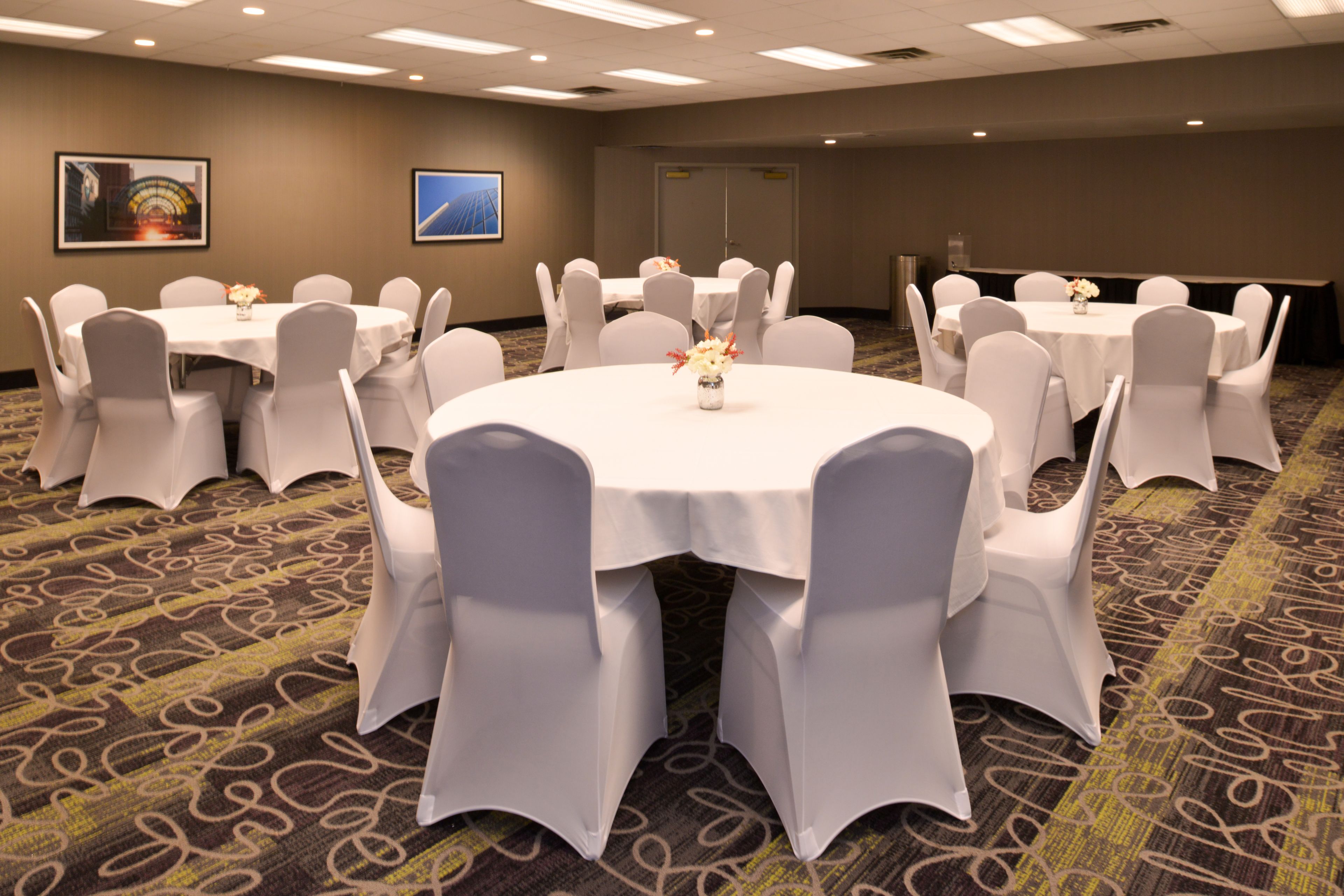 Une salle de conférence avec des tables rondes et des chaises blanches, un sol recouvert de moquette et des tableaux encadrés accrochés aux murs.