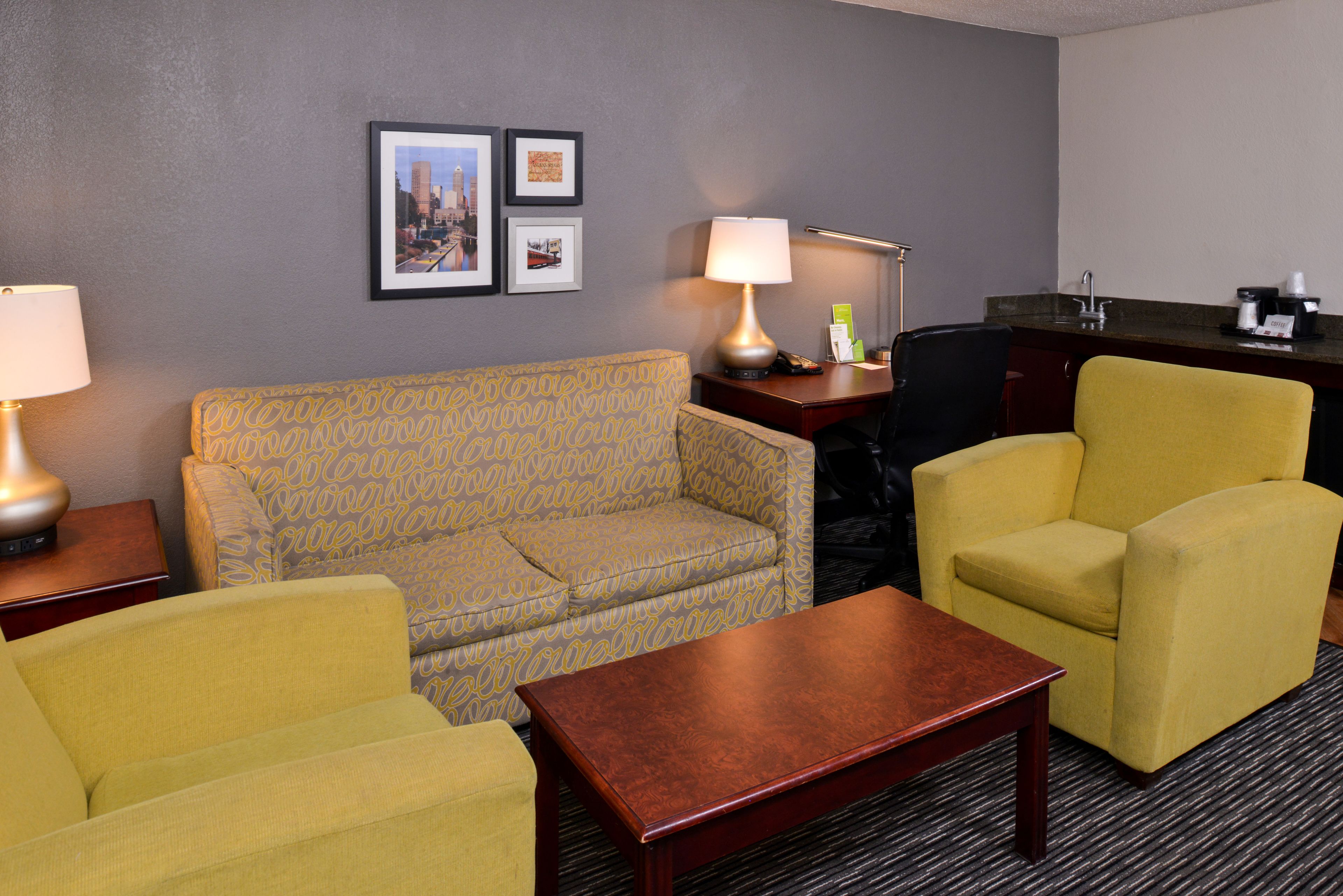 A hotel room with a sofa, two armchairs, a coffee table, a desk with a lamp, a framed cityscape picture, and a kitchenette area.