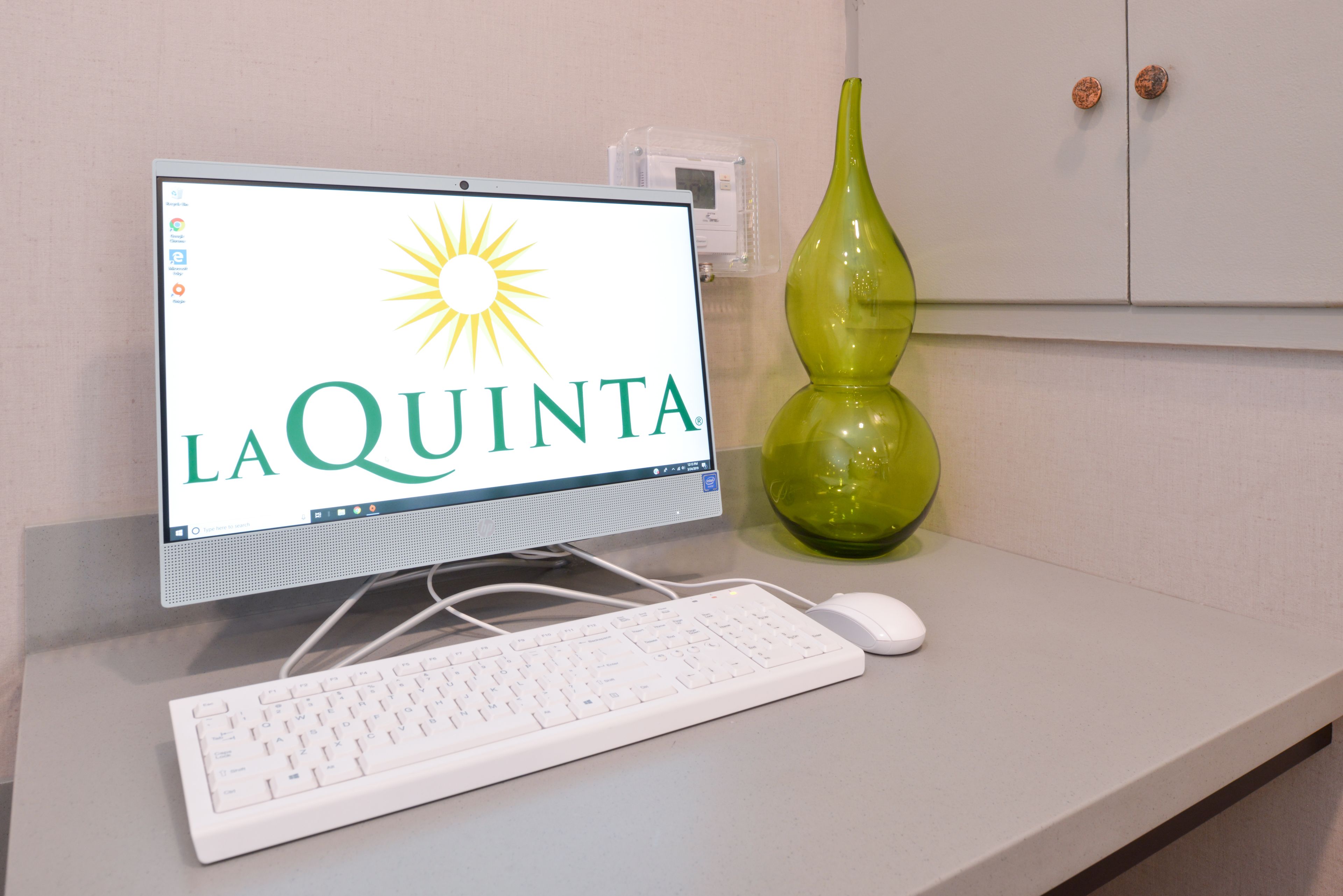 A computer monitor displaying "LA QUINTA" with a sun logo, a white keyboard, a white mouse, and a green vase on a white counter.