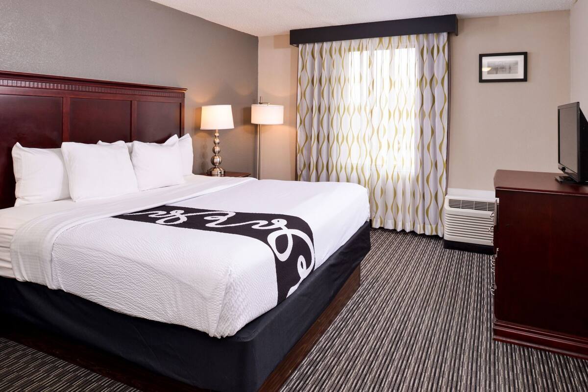 Ein Hotelzimmer mit einem großen Bett, zwei Nachttischlampen, einem Flachbildfernseher, einem Schreibtisch und einem fenster mit Vorhängen.