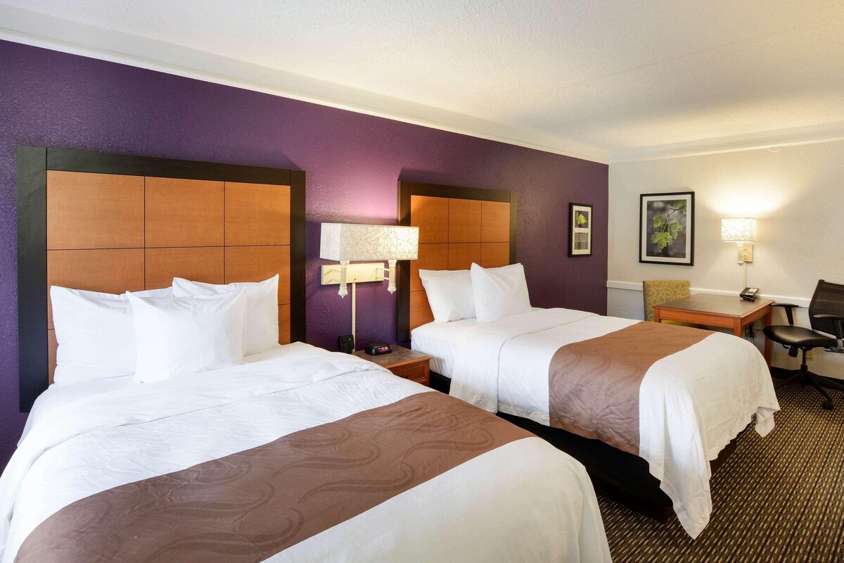 A hotel room with two beds, a nightstand, a lamp, a chair, and a framed picture on the wall.