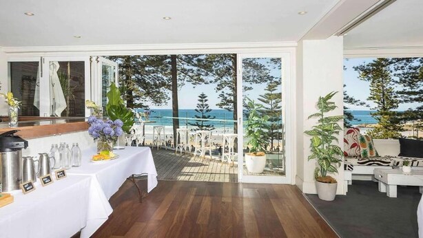 Um quarto amplo com piso de madeira, sala de jantar com mesa branca posta para refeição e área de estar com vista para o mar.