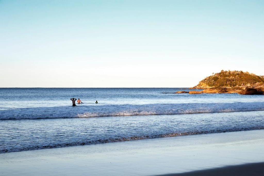 Una playa con aguas tranquilas, una orilla arenosa y una colina rocosa al fondo.