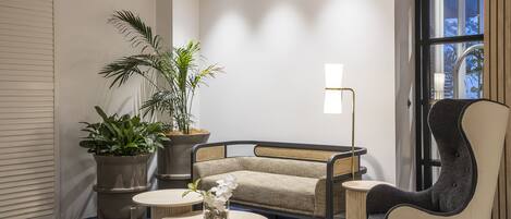A modern lounge area with a sofa, armchair, and round tables, featuring potted plants and a large window.