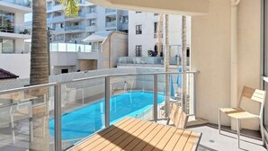 Varanda com vista para a piscina, cadeiras de descanso de madeira e guarda-corpo de vidro.
