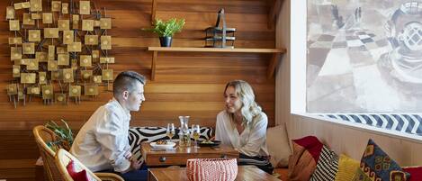 A cozy room with a wooden wall, a checkered floor, and a large window. There is a table with a vase and a plant.