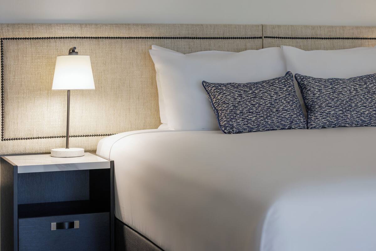 A neatly made bed with white linens and two patterned pillows, a bedside lamp, and a small drawer unit.