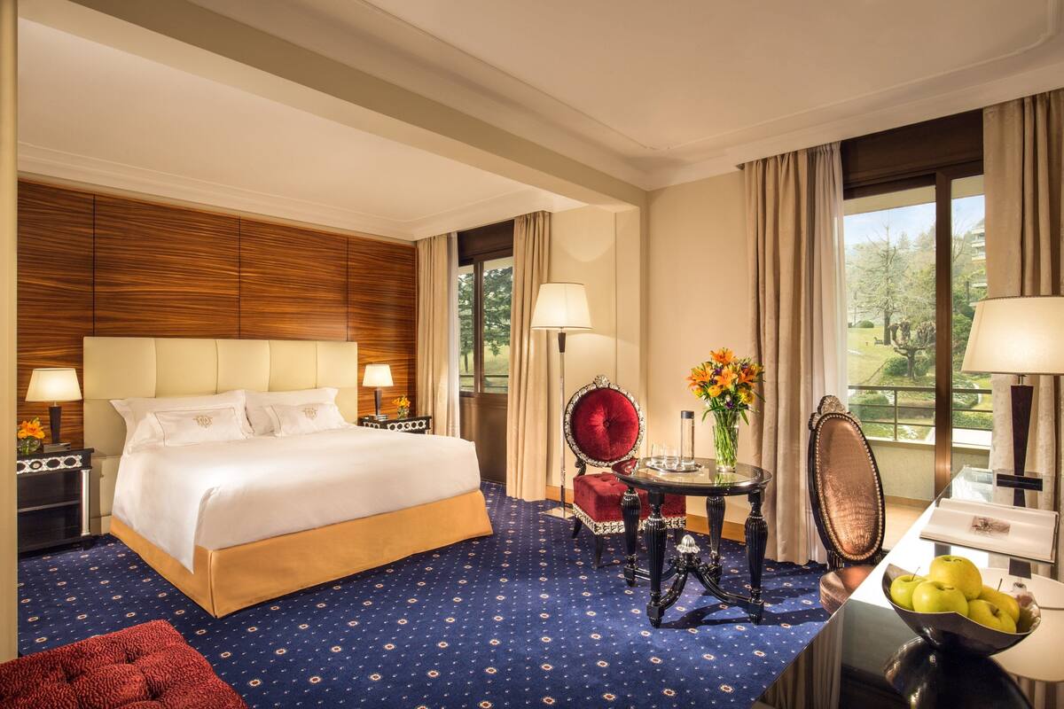 Habitación de hotel con una cama grande, una silla roja, una mesita con un florero y vistas al exterior a través de las ventanas.