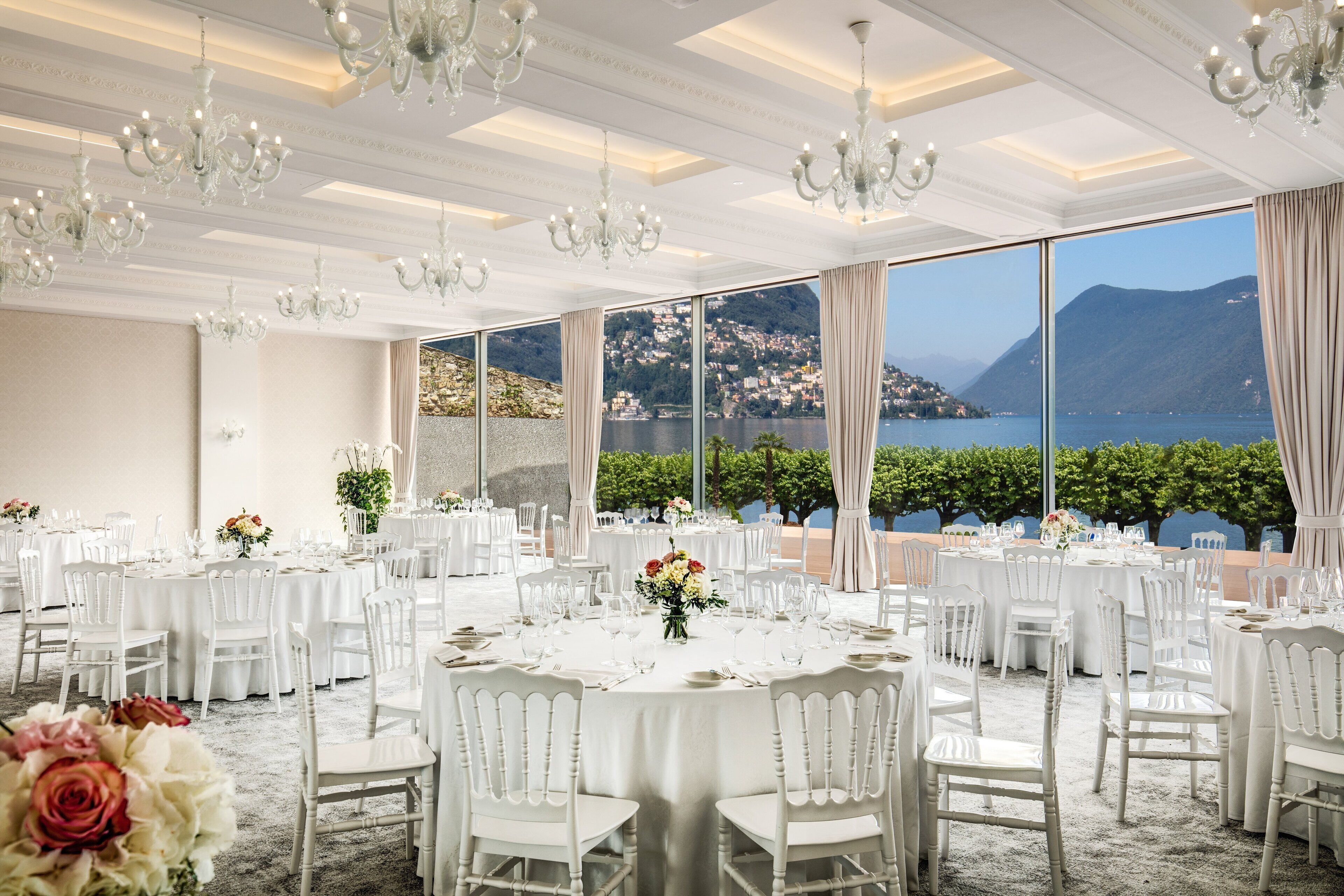 A banquet hall with round tables set for dining, large windows with a view of a lake and mountains, and chandeliers.