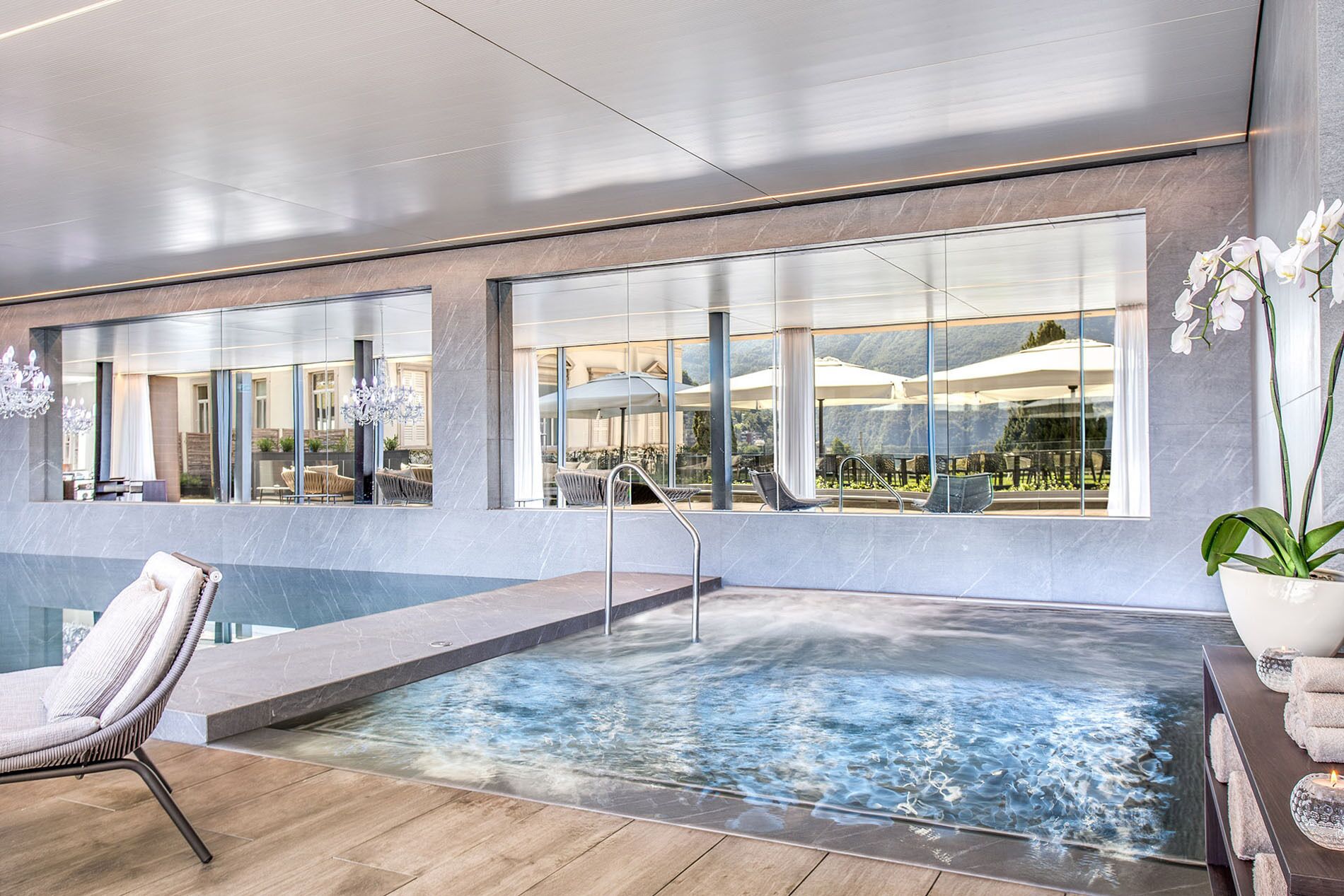Un espace piscine intérieur moderne, doté de parois en verre transparent, d’un fauteuil de relaxation et d’une vue sur la terrasse extérieure.