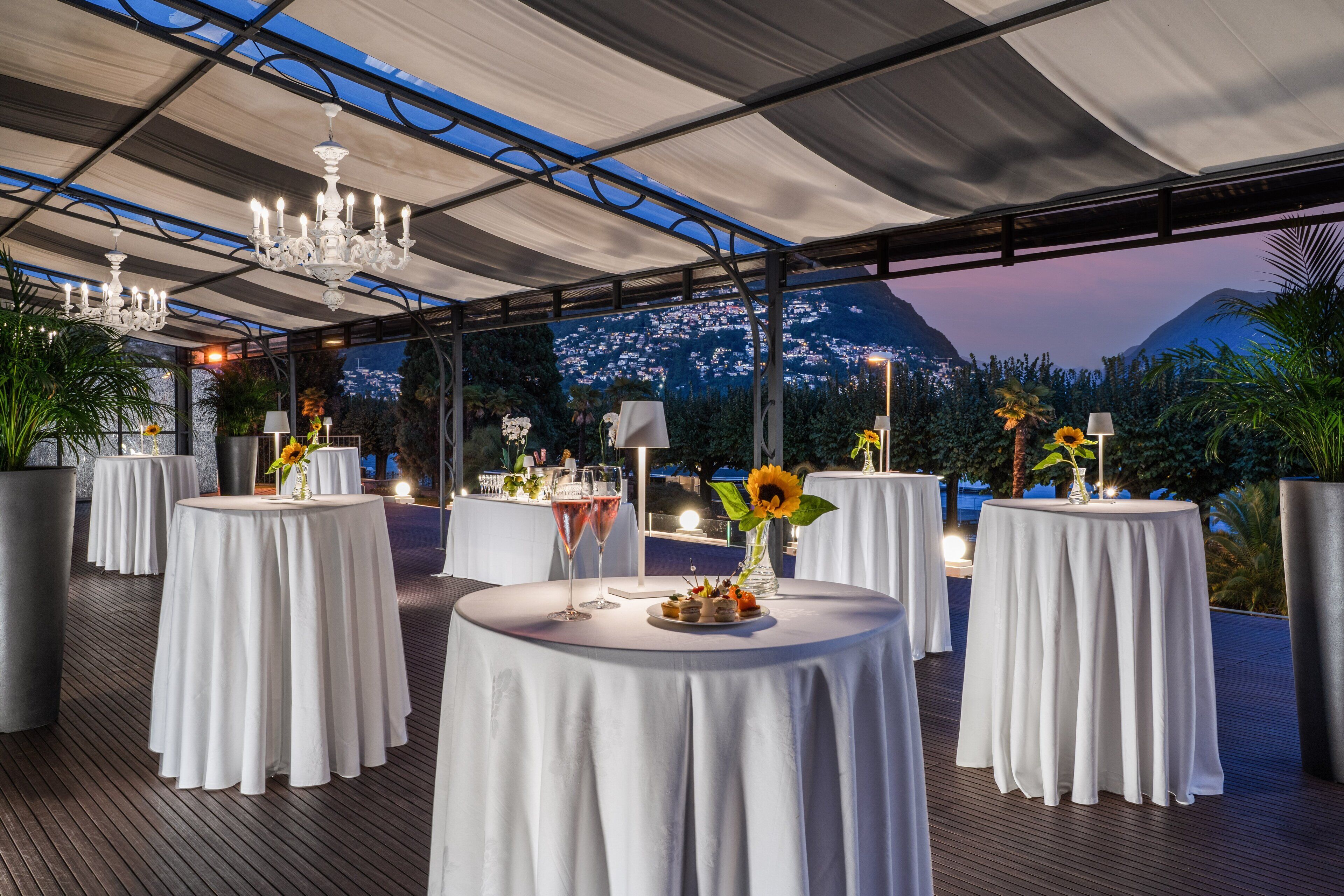 A rooftop dining area with round tables covered in white tablecloths, each with a candle and a flower centerpiece.