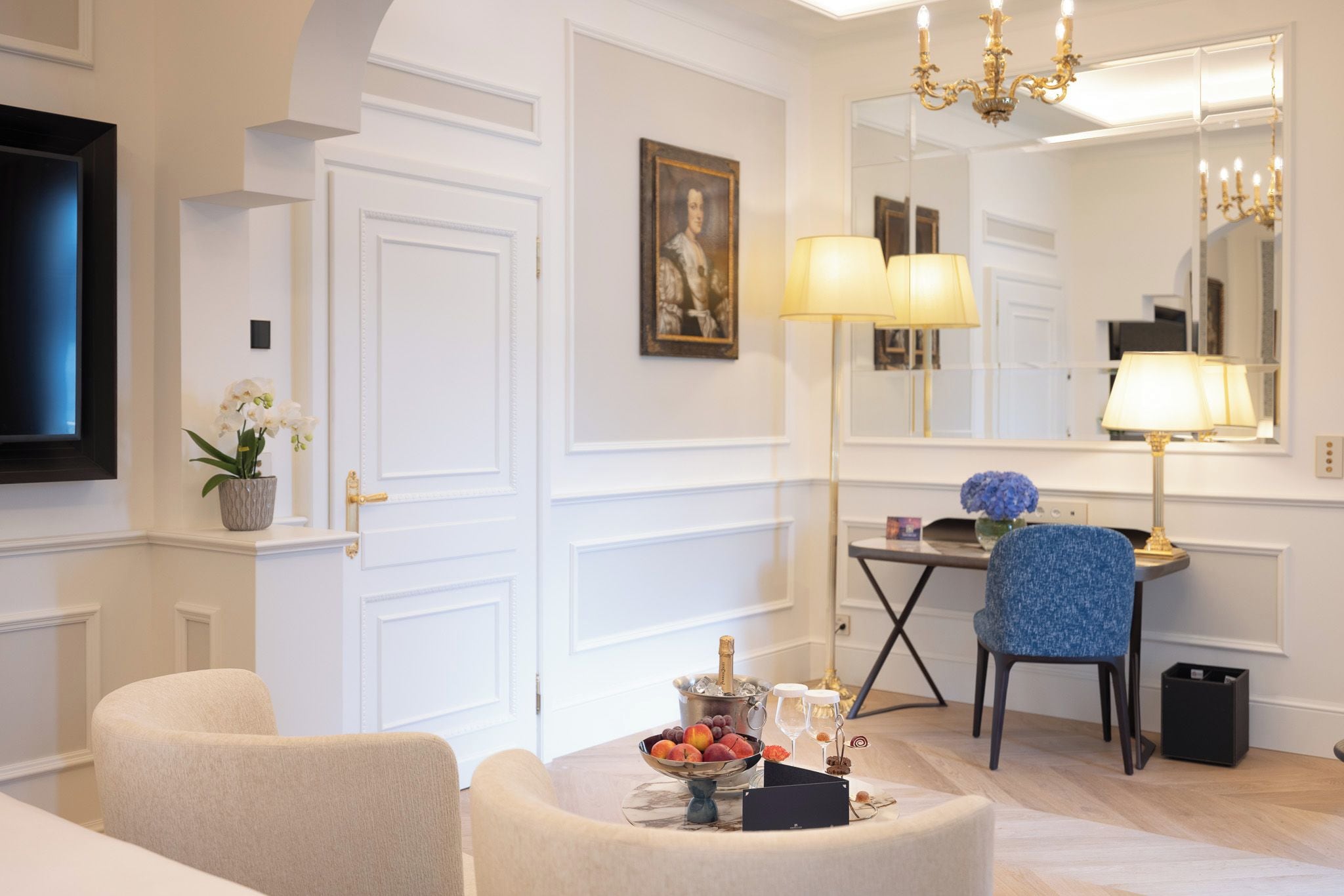 Una habitación bien iluminada con una puerta blanca, un retrato enmarcado, una mesa con fruteros, una botella de champán y una silla azul.