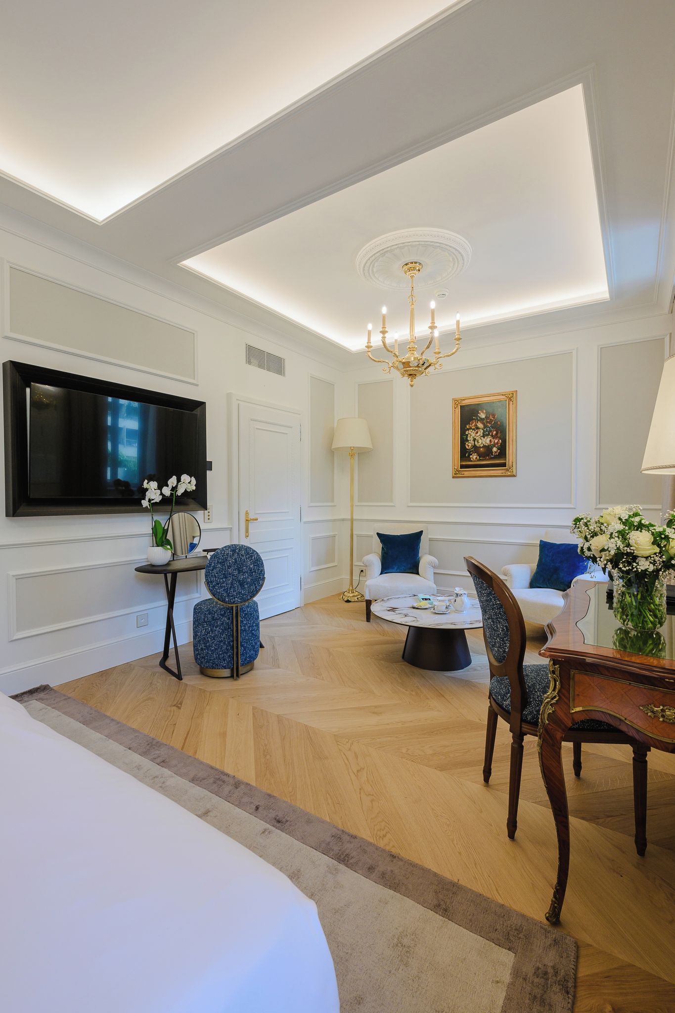 Una habitación de hotel con una cama grande, televisión, candelabro y zona de comedor con sillas y mesa.