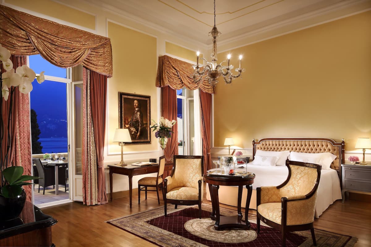 Un dormitorio lujoso con una cama grande, dos sillones, una araña y vistas al exterior.