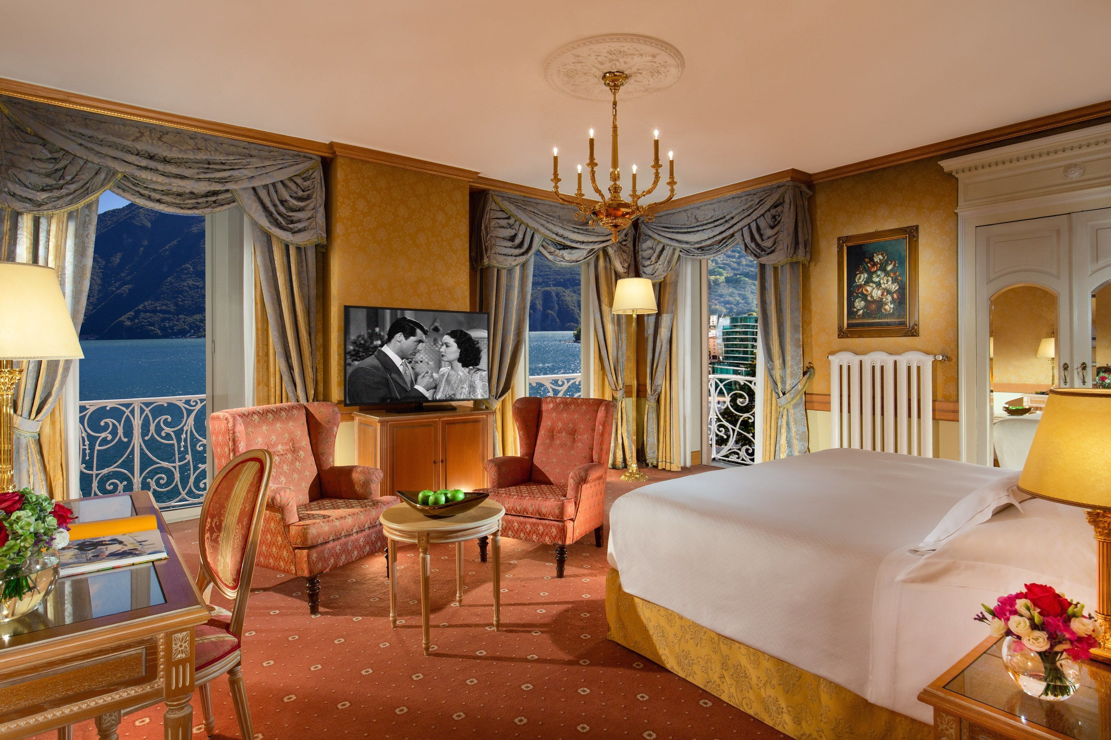 A hotel room with a bed, two armchairs, a small table with a bowl of fruit, a television, and a view of the sea.