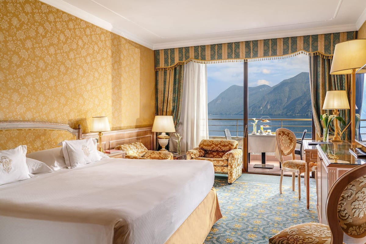 Habitación de hotel con una cama grande, dos sillas, un escritorio y vistas a un lago y montañas.