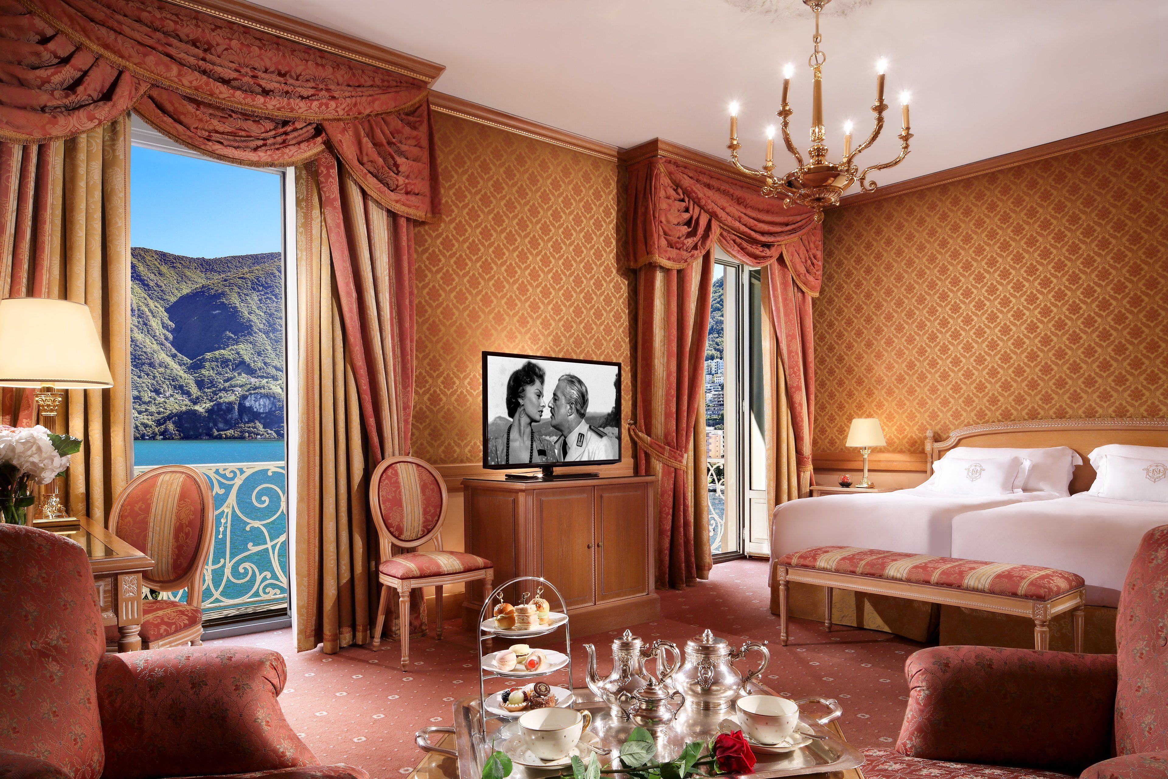 A luxurious hotel room with a large bed, a television, a dining table set with tea and pastries, and a scenic view of a mountainous landscape through the window.