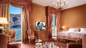 A luxurious hotel room with a large bed, a television, a dining table set with tea and pastries, and a scenic view of a mountainous landscape through the window.