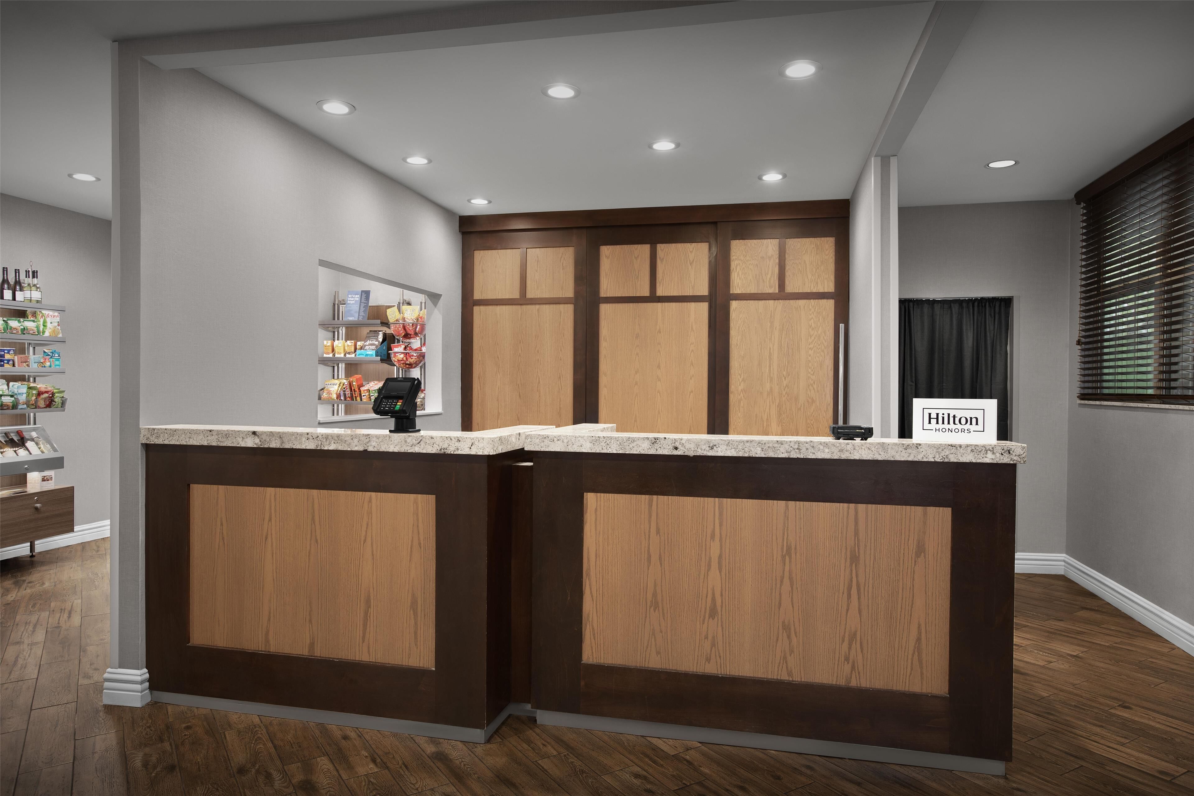 Un espace d’accueil d’hôtel avec un comptoir de réception en bois, un minibar et un panneau indiquant l’adhésion au programme Hilton Honors.