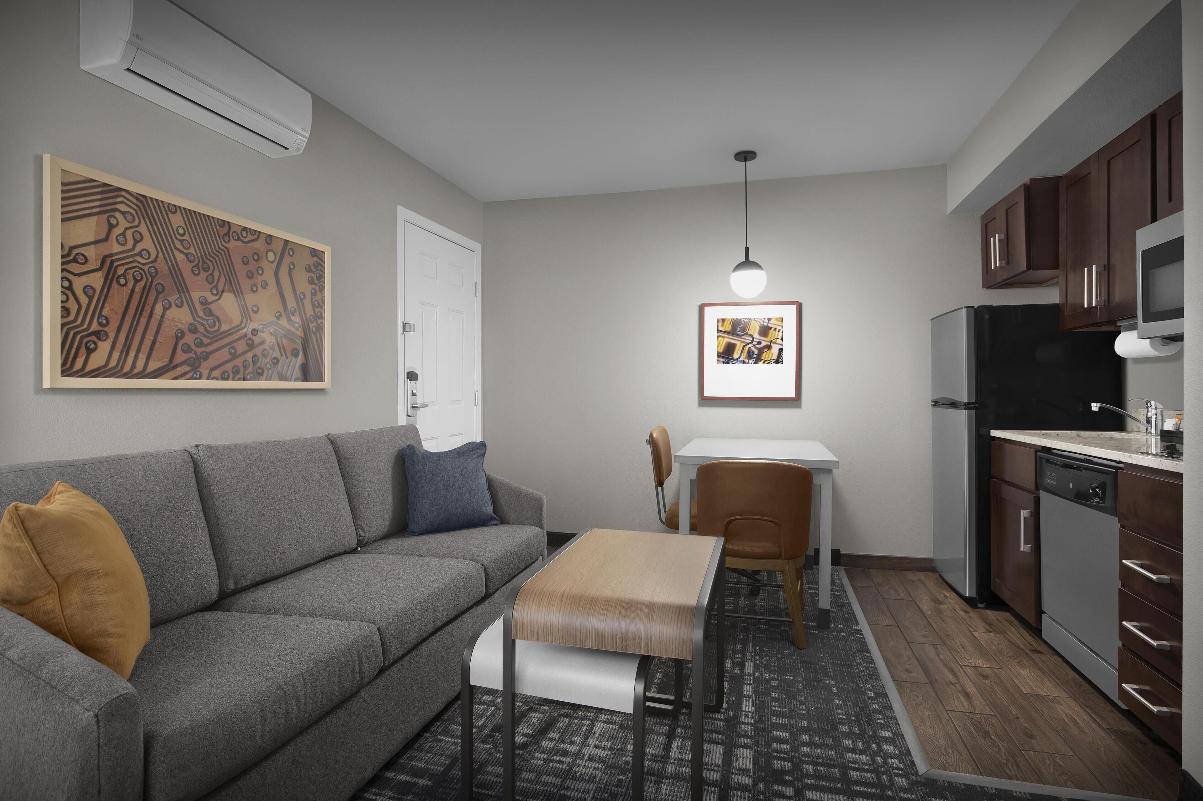 A compact living space with a grey sofa, a small dining table, a kitchen area with a refrigerator and microwave, and artwork on the walls.