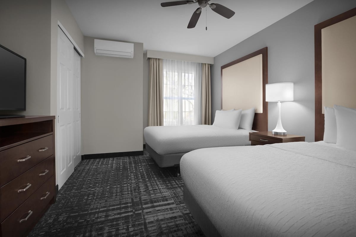 A hotel room with two beds, a ceiling fan, a TV, a dresser, a lamp, and an air conditioning unit.
