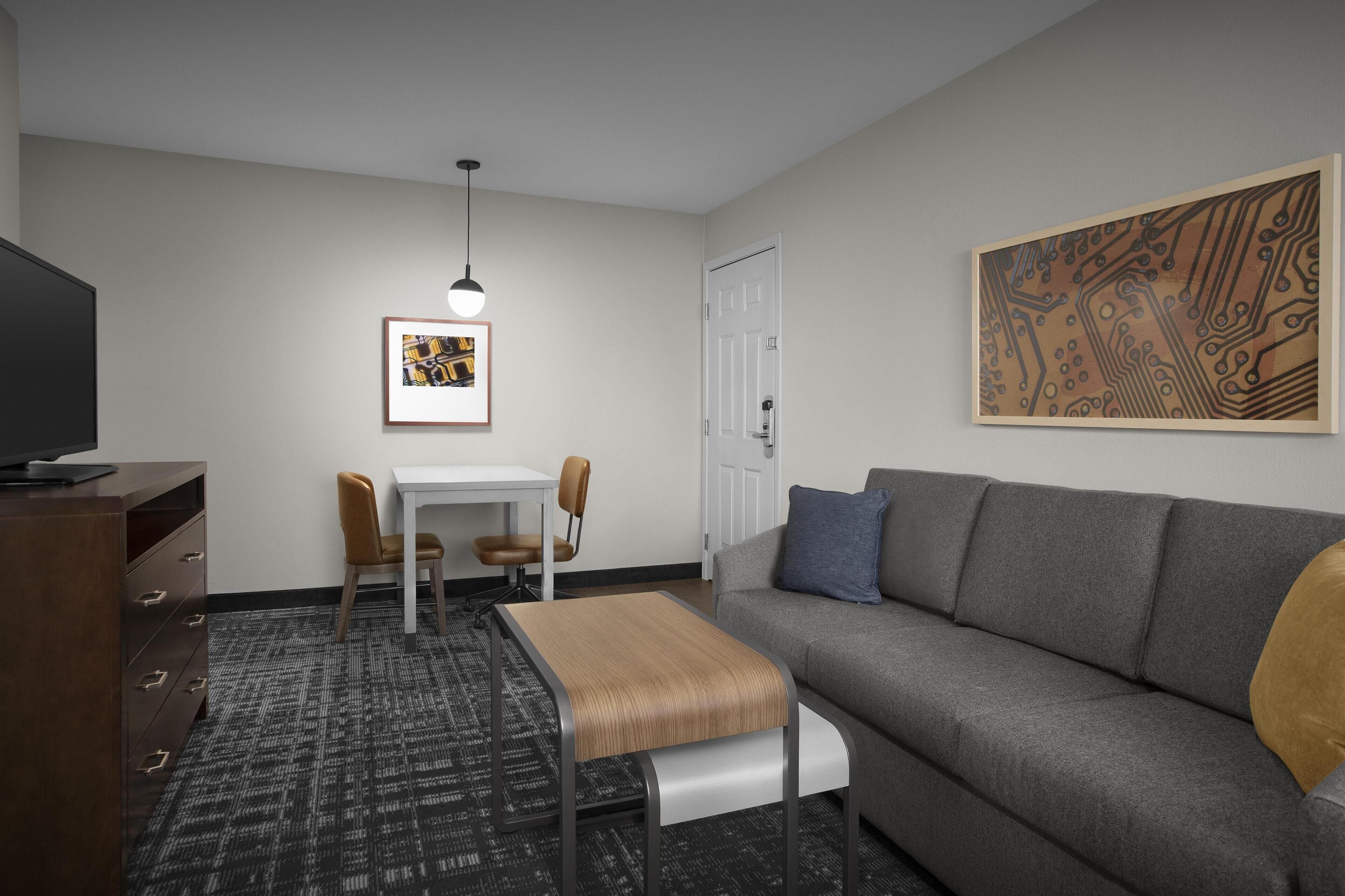 A modern hotel room with a gray sofa, a small wooden table, a single chair, a TV, and a framed artwork on the wall.