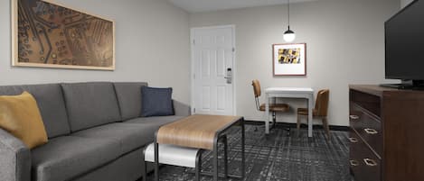 A compact hotel room with a gray sofa, a small wooden table, a white door, and a television.