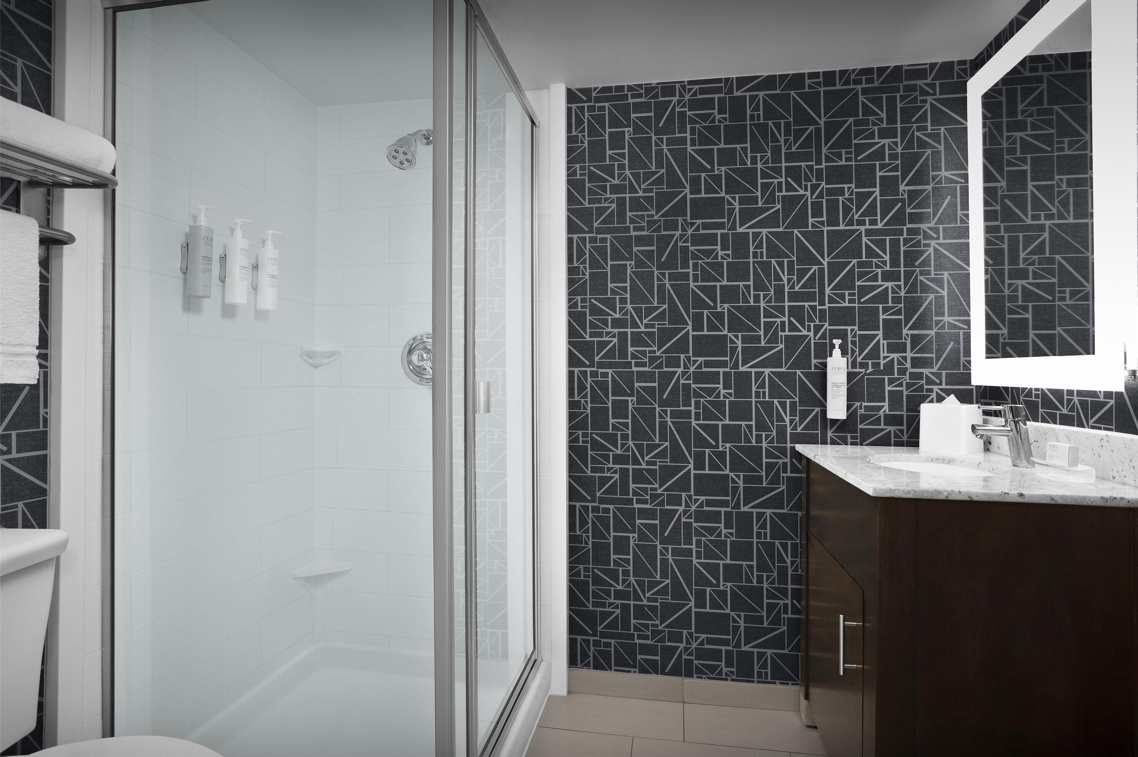 A modern bathroom with a glass-enclosed shower, a dark patterned wall, and a dark wooden vanity with a mirror.