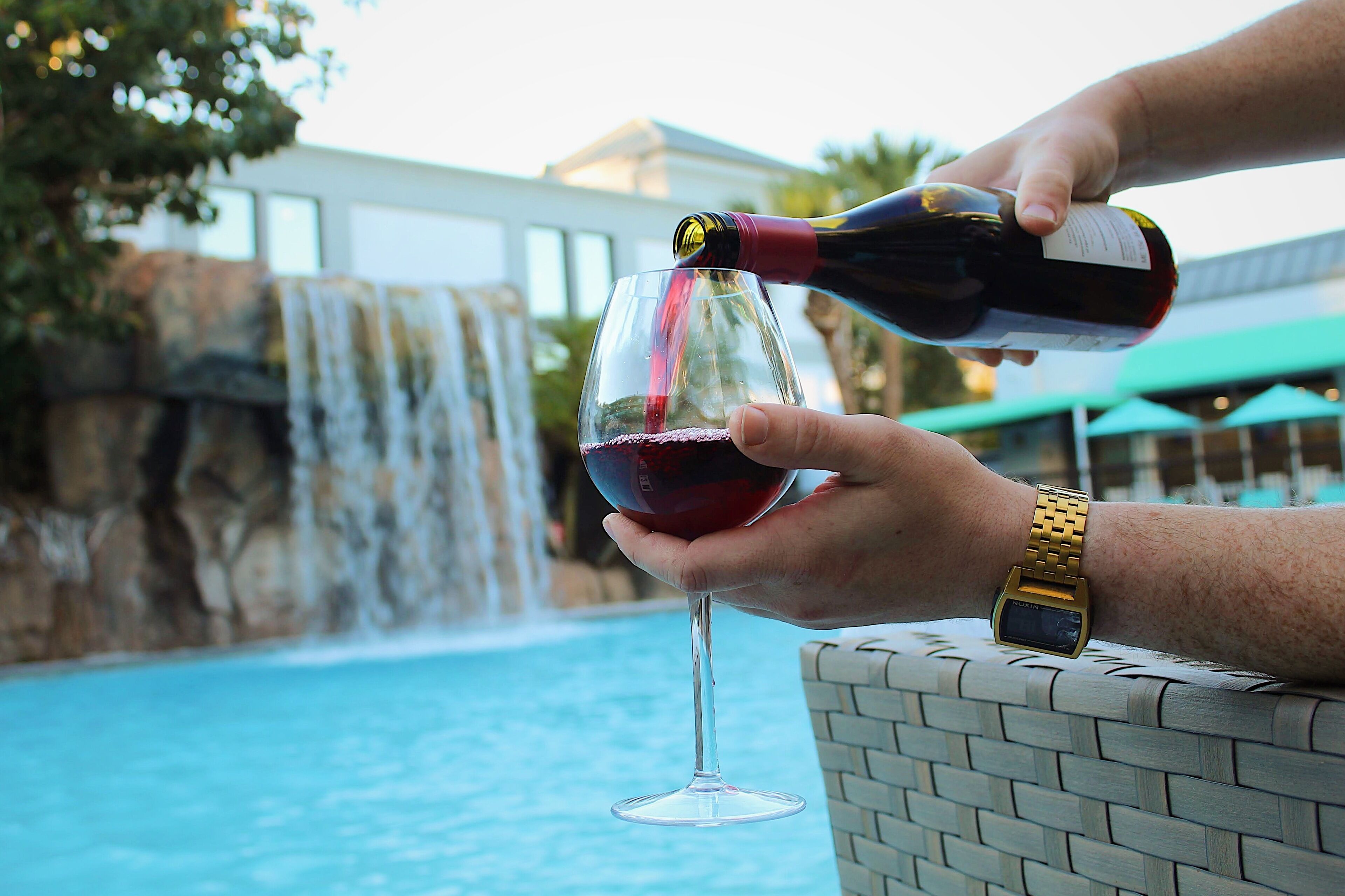 A person pouring red wine into a glass by a pool with a waterfall in the background.