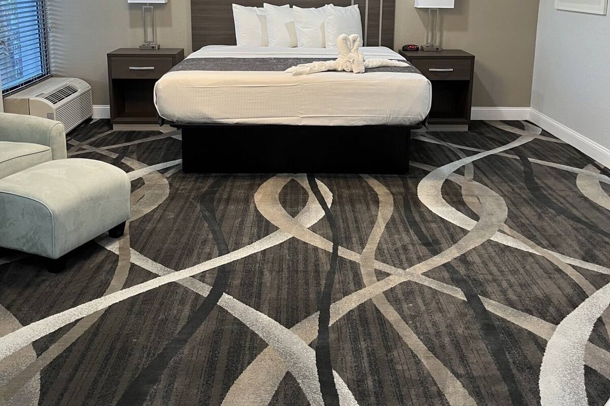 Una habitación de hotel con una cama grande, dos mesitas de noche con lámparas, un candelabro moderno, una alfombra estampada y una ventana con persianas.