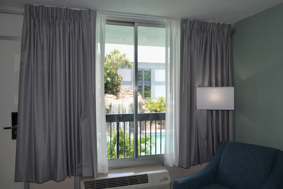 Una habitación con un ventanal grande, cortinas grises, un sillón azul y una lámpara de pared.