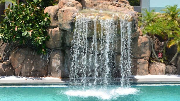 A clear blue pool with a waterfall feature surrounded by rocks and greenery. - Opal Hotel & Suites (Altamonte Springs)