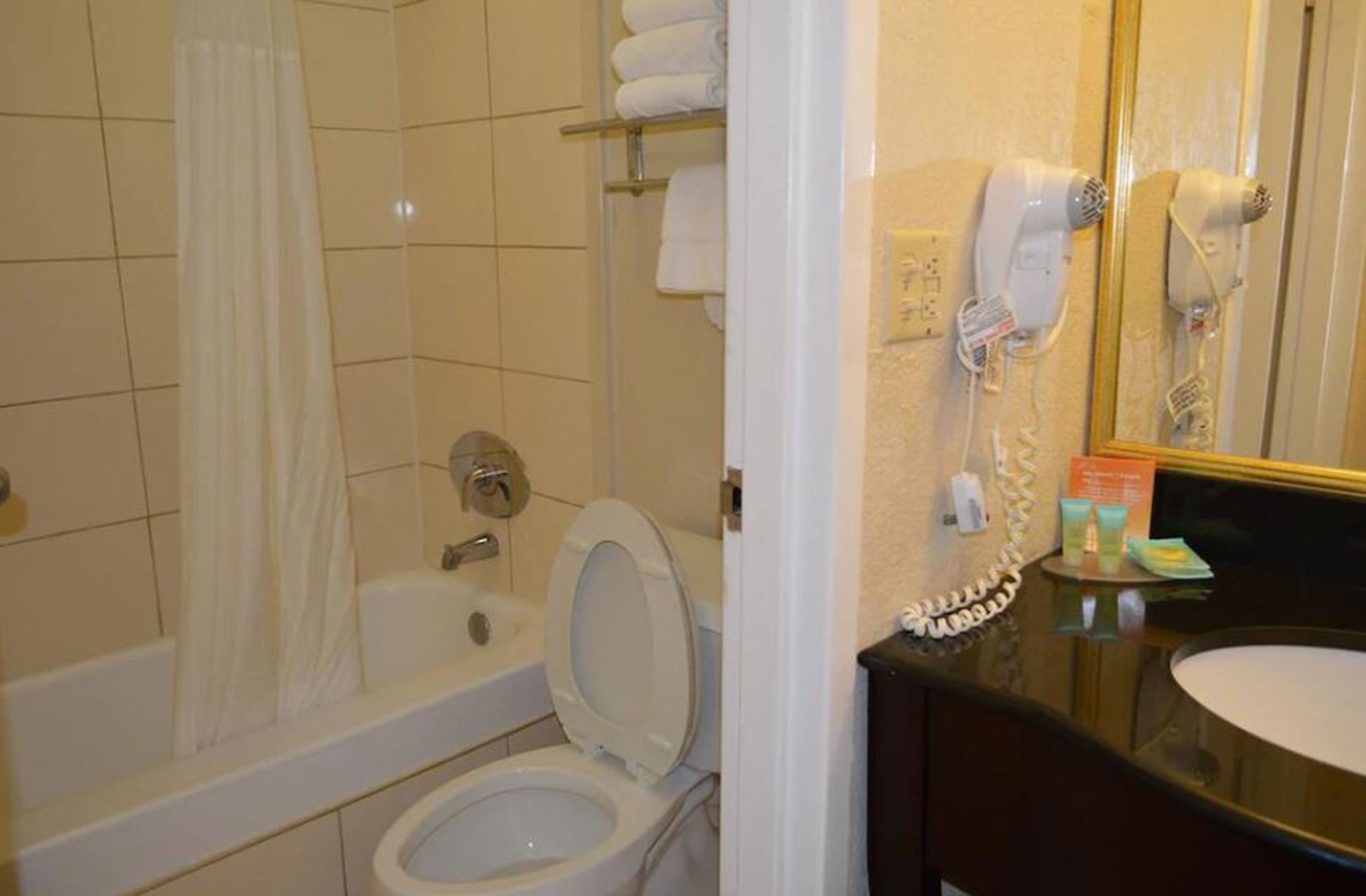 Un baño con bañera, inodoro y lavamanos con espejo. Hay secadores de pelo y artículos de aseo en la encimera.