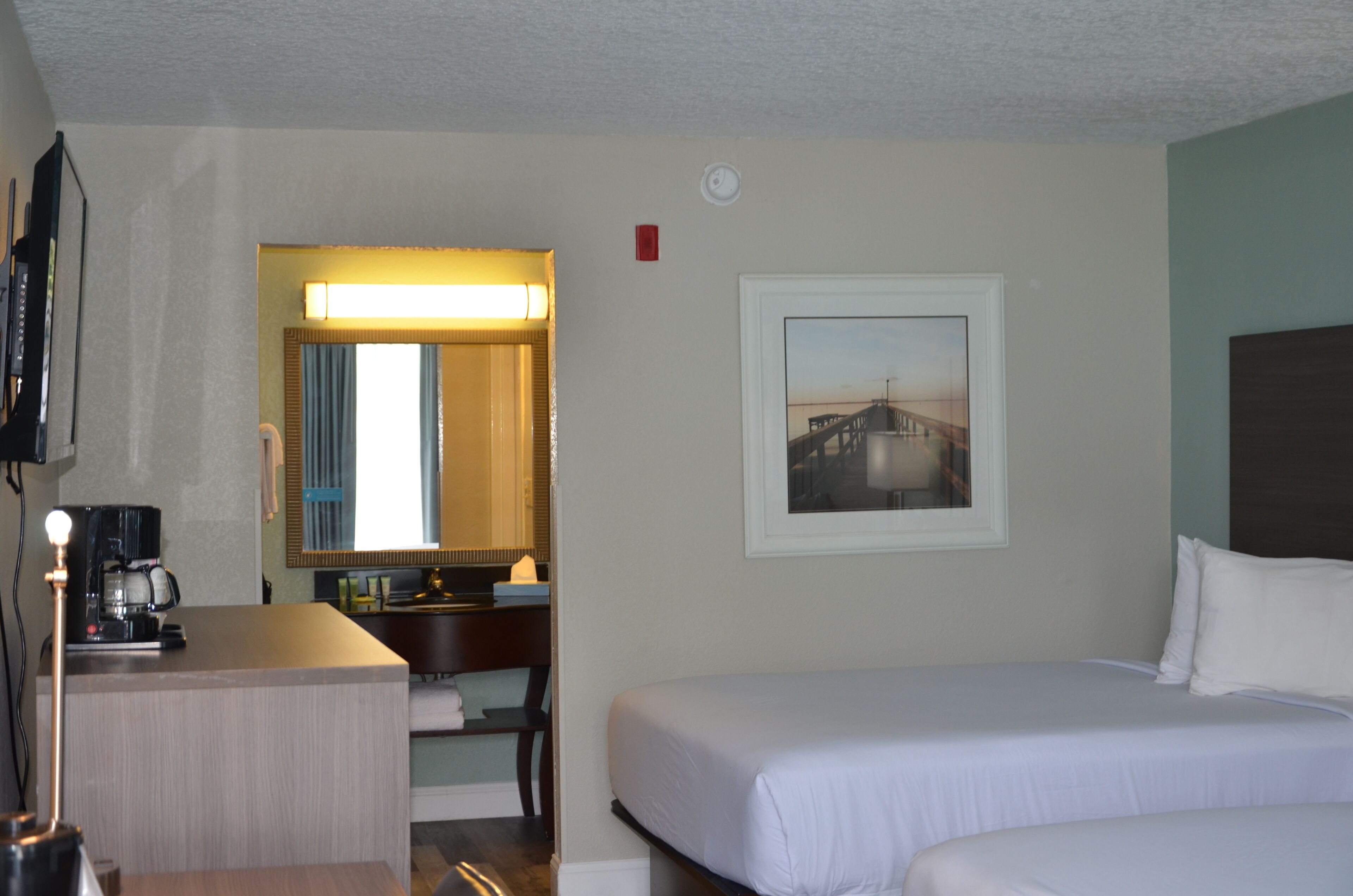 A hotel room with a bed, a desk with a coffee maker, a mirror, and a framed picture on the wall.