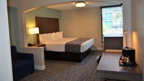 A hotel room with a bed, bedside table, lamp, chair, desk, coffee maker, and a window with blinds. - Opal Hotel & Suites (Altamonte Springs)