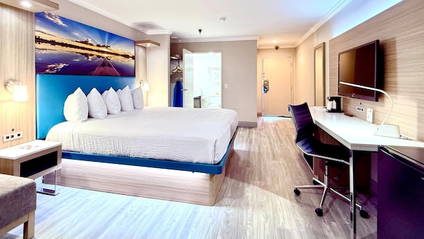 Une chambre d’hôtel moderne dotée d’un grand lit, d’un bureau, d’une télévision à écran plat et d’un tableau représentant un coucher de soleil sur l’eau.