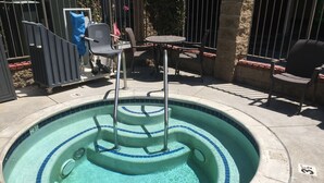 Un bain à remous circulaire, bordé de carreaux bleus, avec une échelle en métal et une petite table entourée de chaises.