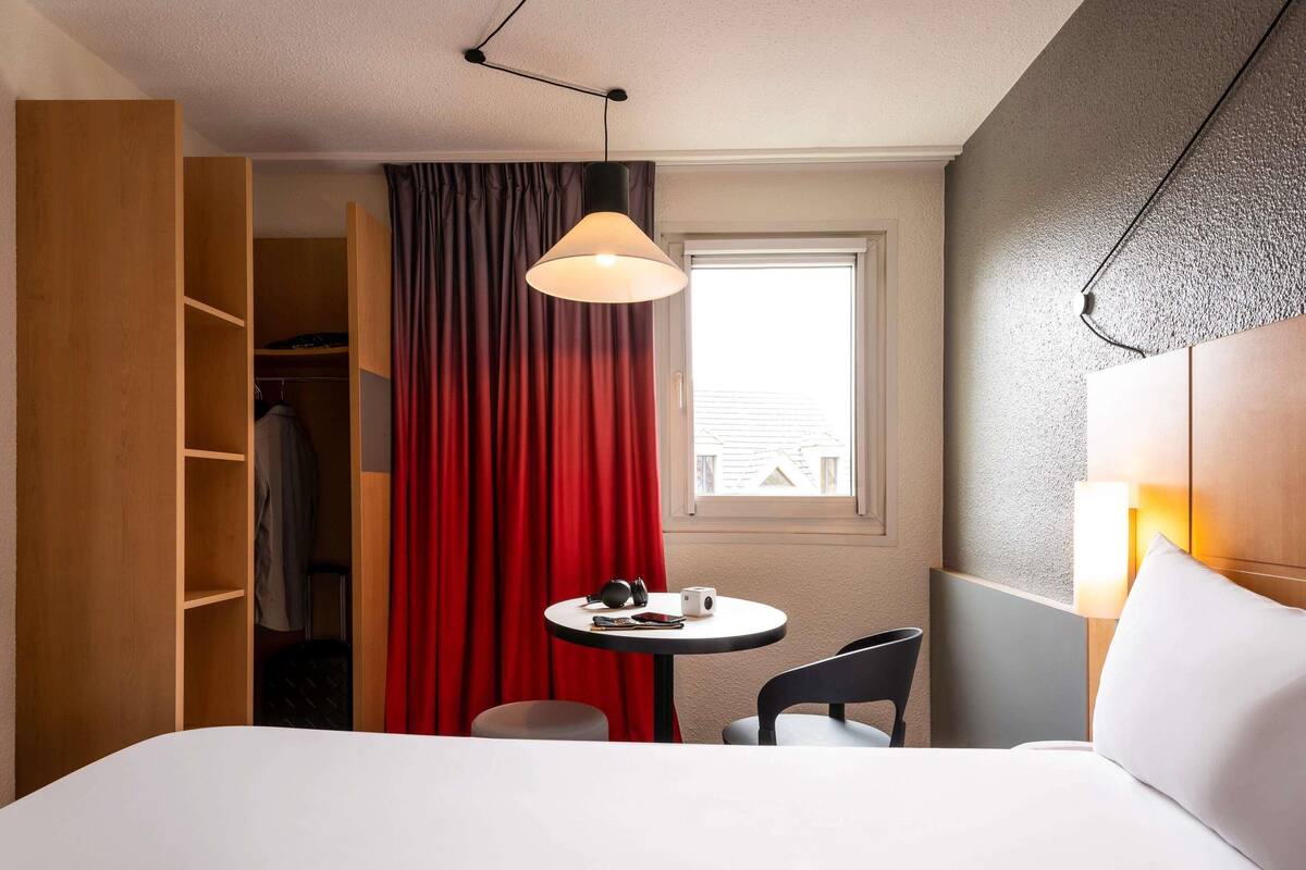 Een hotelkamer met een bed, een ronde tafel met stoel, een kledingkast, een raam met gordijnen en een hanglamp.