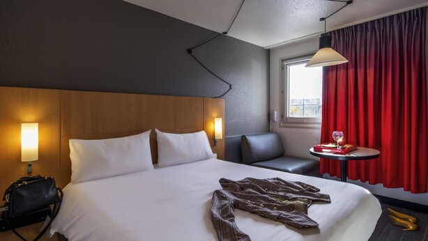 Una habitación de hotel con una cama grande, una lámpara de noche, una mesita con un vaso y una cortina roja.