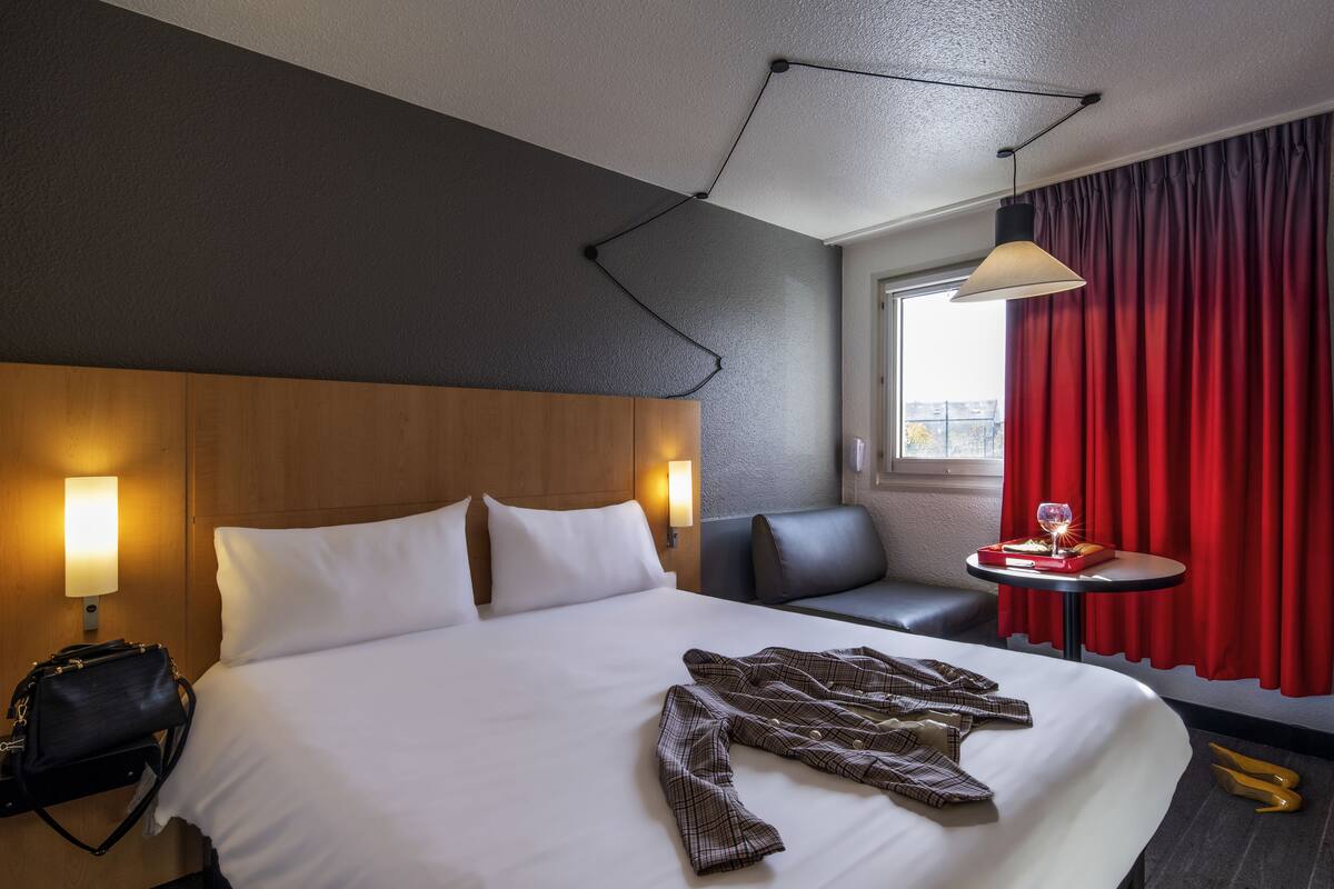 Een hotelkamer met een groot bed, een bedlampje, een klein tafeltje met een glas en een rood gordijn.
