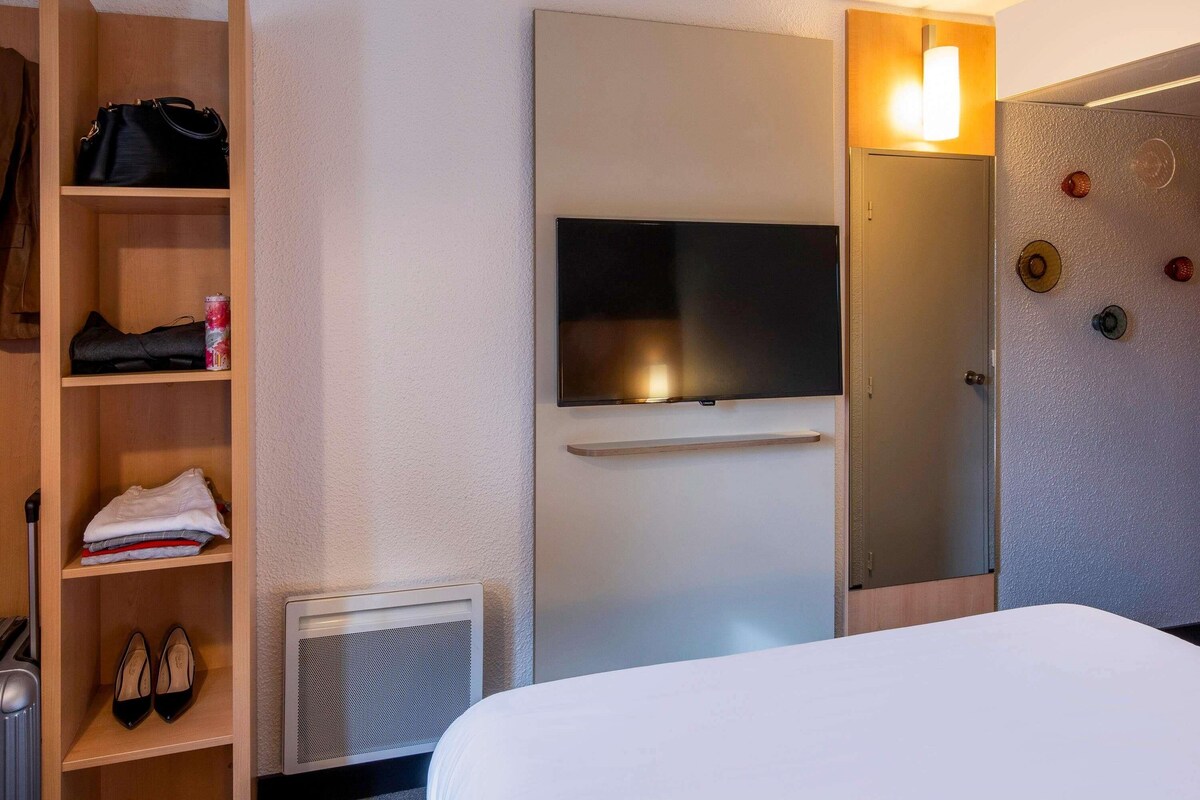 A hotel room with a bed, a television mounted on the wall, a wardrobe, and a shoe rack.