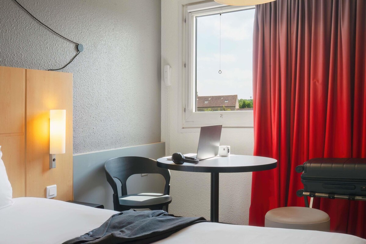 Een hotelkamer met een bed, een bureau met een laptop, een stoel en een rood gordijn.