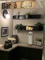Zona de bar con un letrero que dice "Bar à Eau", una encimera blanca, una máquina de café, un teléfono y tres botellas de sirope Monin.