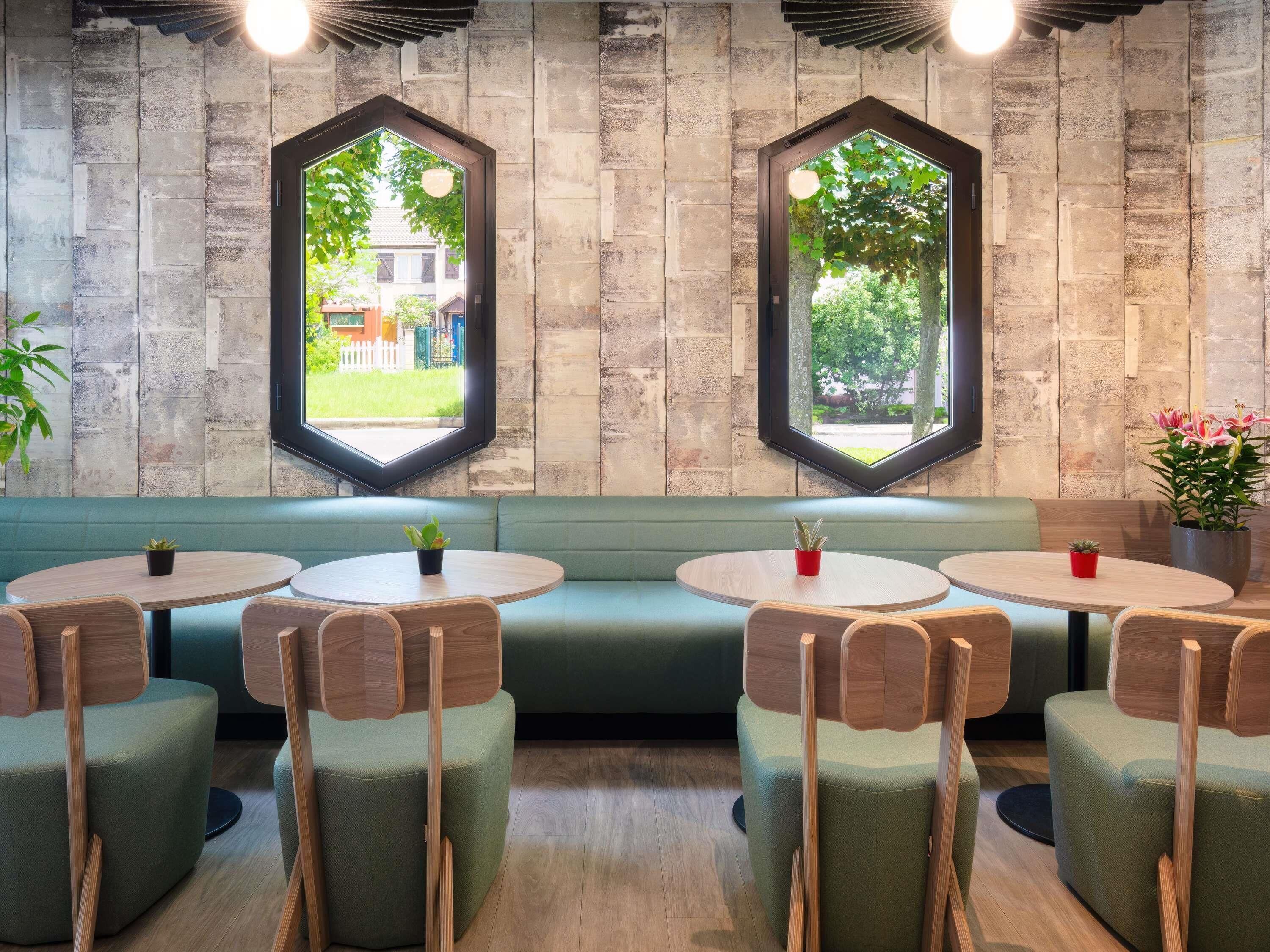 A restaurant interior with a green booth seating area, round tables, and hexagonal windows with views of greenery.